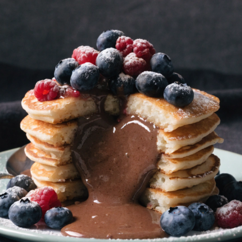 Pile de pancakes moelleux nappés de sauce chocolat type Nutella avec fruits rouges et sucre glace.
